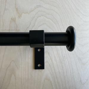 May include: Black metal curtain rod with a round finial and a square bracket. The rod is mounted on a light wood surface.