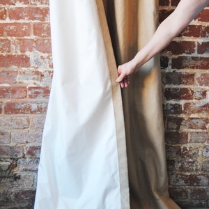 May include: A pair of curtains, one white and one beige, are partially open. The white curtain is pulled back to reveal the beige curtain behind it. The curtains are hanging in front of a brick wall.