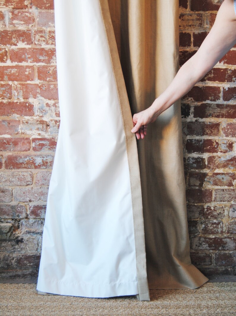 May include: A pair of curtains, one white and one beige, are partially open. The white curtain is pulled back to reveal the beige curtain behind it. The curtains are hanging in front of a brick wall.
