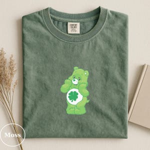 May include: Moss green t-shirt featuring a green Care Bear with a four-leaf clover on its belly. The shirt has a classic crew neck and a tag that reads "Comfort Colors". The shirt is folded neatly on a neutral surface.