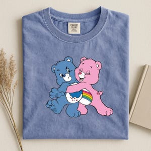 May include: A blue t-shirt featuring a graphic of two cartoon bears hugging. One bear is blue and the other is pink, with a rainbow and cloud design between them. The shirt has a Comfort Colors tag.