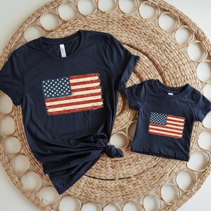 May include: Two navy blue t-shirts with a distressed American flag design. The larger shirt is adult-sized, and the smaller one is for a child. The flag features red and white stripes and a blue field with white stars. The shirts are displayed on a woven, circular mat.