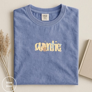 May include: Blue jean Comfort Colors t-shirt with the word "auntie" in a yellow gingham pattern with a cartoon bear holding a honey pot. The shirt has a crew neck and a tag on the collar.