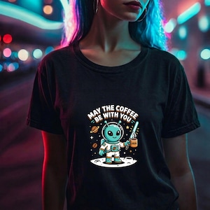 May include: Black t-shirt featuring a cartoon alien holding a coffee mug with the word "JAVA". The alien is standing on a moon-like surface with planets and stars in the background. The text "MAY THE COFFEE BE WITH YOU" is above the alien.