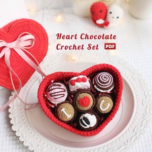 May include: A red crochet heart-shaped box filled with crochet chocolates. The chocolates have various designs, including swirls, stripes, and hearts. The box sits on a white plate, and the text "Heart Chocolate Crochet Set PDF" is visible.