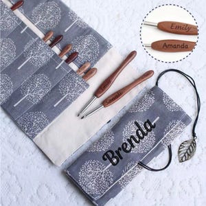 May include: A gray fabric crochet hook roll with a white tree pattern, featuring personalized wooden hooks labeled with names like Emily, Amanda, and Sonali. The roll is embroidered with the name Brenda and includes a metal leaf charm.