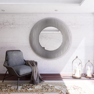 Op de afbeelding: Een modern interieur met een grote, ronde spiegel met een getextureerde, geribbelde lijst. Een grijze fauteuil met bijpassende deken staat op een patroonkleed. Twee decoratieve kandelaars completeren de sfeer.