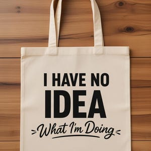 May include: Cream-colored tote bag with black text that reads "I HAVE NO IDEA >What I'm Doing<". The bag has two handles and is made of a sturdy fabric. The background is a wooden surface.