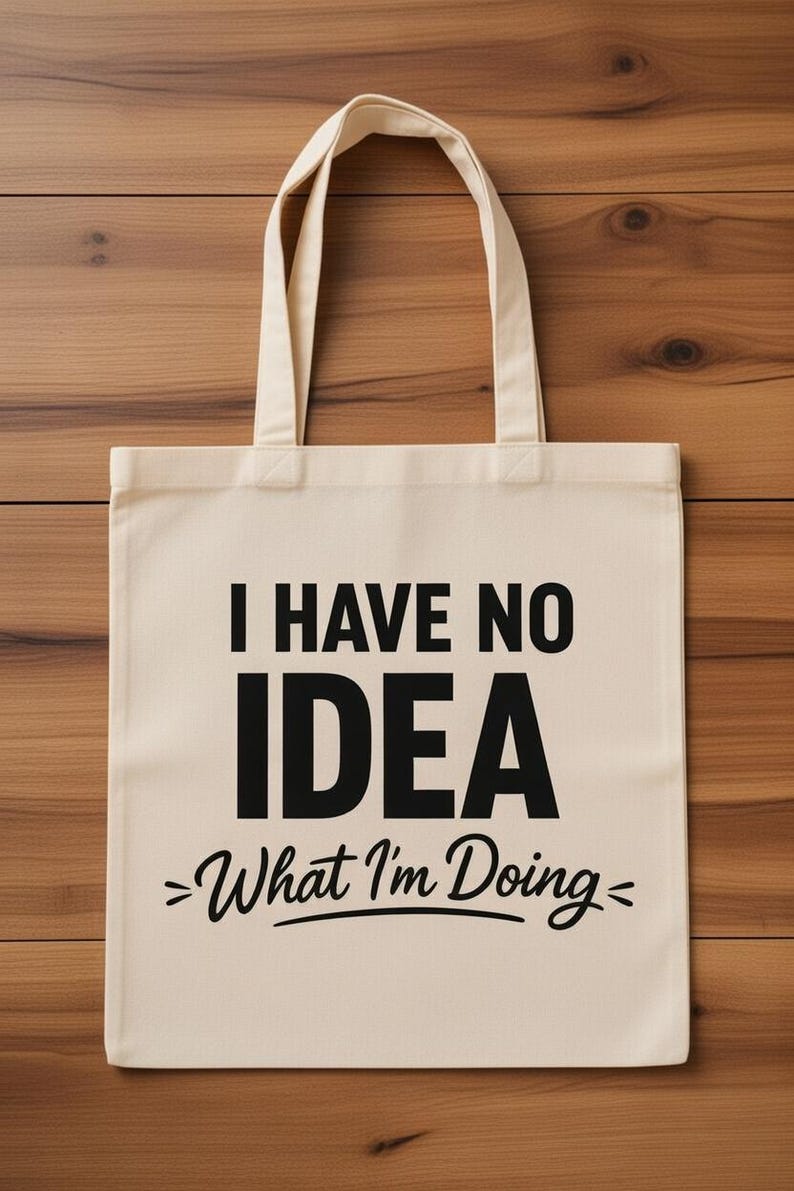 May include: Cream-colored tote bag with black text that reads "I HAVE NO IDEA >What I'm Doing<". The bag has two handles and is made of a sturdy fabric. The background is a wooden surface.