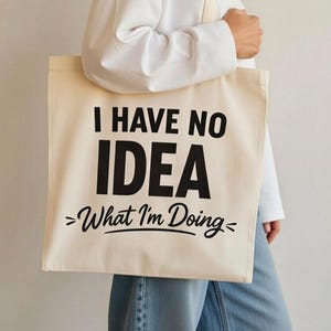 May include: Cream-colored tote bag with the text "I HAVE NO IDEA >What I'm Doing<". The bag has a rectangular shape and long straps. The text is in a bold, black font.