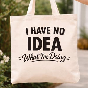 May include: A beige canvas tote bag with black text that reads "I HAVE NO IDEA - What I'm Doing-". The bag is held up by a person. The background is blurred with greenery.