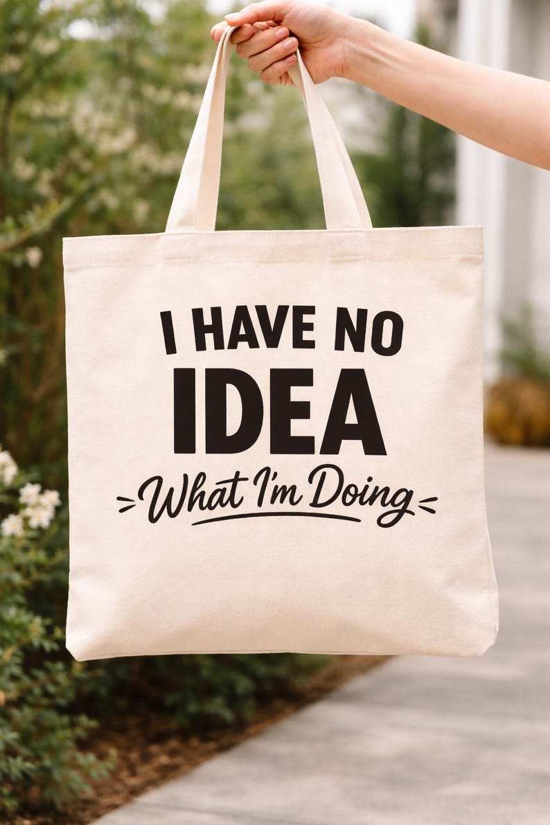 May include: A beige canvas tote bag with black text that reads "I HAVE NO IDEA - What I'm Doing-". The bag is held up by a person. The background is blurred with greenery.