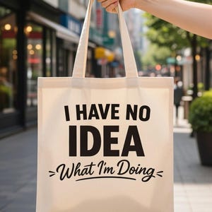 May include: A beige canvas tote bag with black text that reads "I HAVE NO IDEA >What I'm Doing<". The bag is held up in front of a blurred city background.