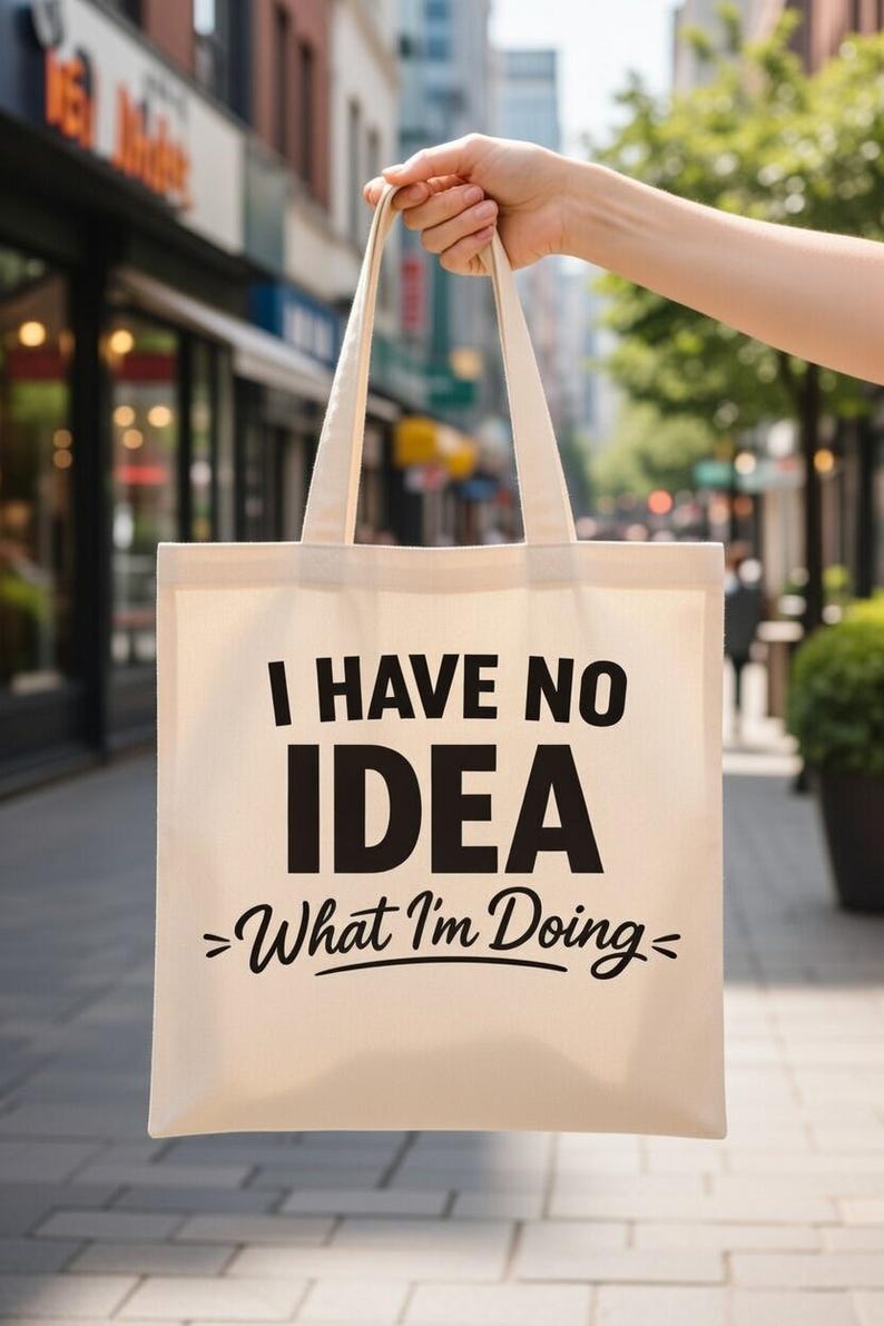 May include: A beige canvas tote bag with black text that reads "I HAVE NO IDEA >What I'm Doing<". The bag is held up in front of a blurred city background.