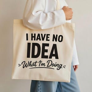 May include: Cream-colored tote bag with the text "I HAVE NO IDEA >What I'm Doing<". The bag has a simple design with black lettering and a neutral color scheme. The bag is carried by a person.