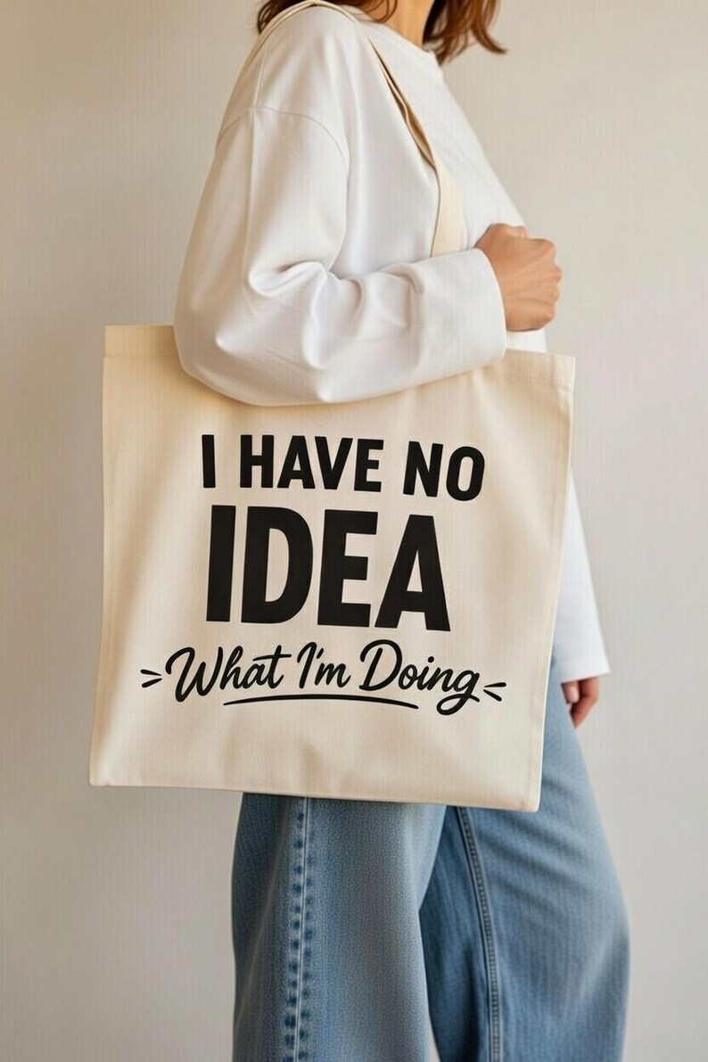 May include: Cream-colored tote bag with the text "I HAVE NO IDEA >What I'm Doing<". The bag has a simple design with black lettering and a neutral color scheme. The bag is carried by a person.
