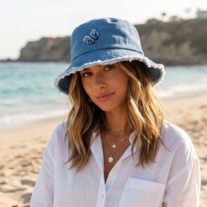 Puede incluir: Un sombrero de pescador de mezclilla azul claro con borde deshilachado y un pequeño gráfico de ojo bordado en la parte delantera. Se combina con una camisa blanca de manga larga con botones, pantalones cortos de mezclilla azul claro y un bolso de lona beige.