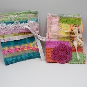May include: Two rectangular fabric journals featuring layered textile strips, lace trim, and decorative stitching. The left journal has teal, pink, and white ribbons with a white bow. The right journal features orange, yellow, and pink patches with a purple doily.