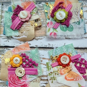 Könnte beinhalten: Vier quadratische Stoffverzierungen aus geschichteten Resten von Blumen- und gemusterten Textilien, rosa Häkelakzenten, Blattgoldfragmenten und weißen Knöpfen. Jedes Stück weist ausgefranste Kanten und dekorative Bänder in verschiedenen Farben auf.