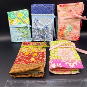 May include: Five rectangular fabric needle cases with ribbon ties, featuring various floral and geometric patterns in green, blue, pink, red, and yellow. A clear plastic ruler is positioned in front, showing a length of approximately 7.5 inches.