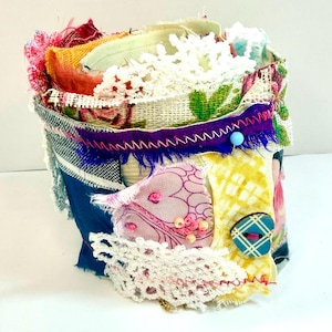 Può includere: Un bracciale a polsino in tessuto fatto a mano con un design patchwork. Il bracciale presenta una varietà di tessuti colorati, tra cui stampe floreali, pizzo e materiali strutturati. È adornato con un bottone blu e uno spillo blu.