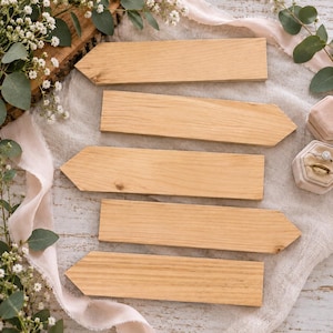 May include: Five light brown wooden signs, each shaped like a rectangular arrow with a pointed end. The wooden planks feature a natural grain pattern and smooth surfaces, arranged in a vertical stack on a textured fabric surface.