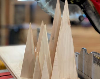 May include: A collection of unfinished wooden mountain-shaped decorations of varying sizes. The light-colored wood pieces are arranged on a wooden surface, with a saw in the background. These decorative items could be used for home decor or craft projects.