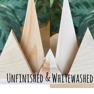 May include: Wooden geometric mountain decorations in various sizes and finishes. Some are unfinished, while others are whitewashed. The text "UNFINISHED & WHITEWASHED" is displayed across the bottom of the image. Green foliage is visible in the background.
