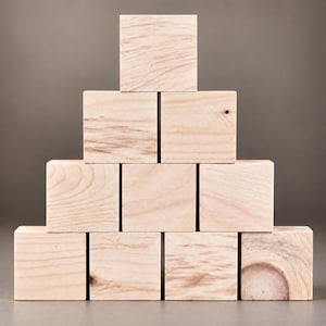 May include: A pyramid stack of ten light-colored wooden blocks arranged in four rows. The blocks feature visible natural wood grain patterns and textures, with one block on the bottom right displaying a prominent circular knot in the wood grain.