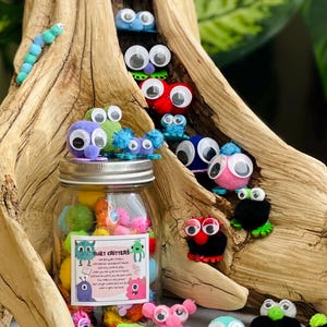 May include: A collection of colorful pom-pom creatures with googly eyes, some nestled in a wooden structure and others in a glass jar. The jar has a label that reads "Quiet Critters". The creatures are in various colors, including blue, pink, green, and black.