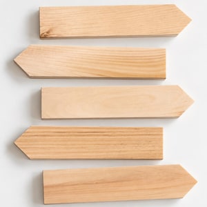 May include: Five light-colored wooden signs, each shaped like a rectangular arrow pointing to the right. The signs feature a natural wood grain pattern with smooth, flat surfaces and clean, straight edges, arranged in a vertical stack.