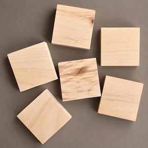 May include: A collection of six square wooden blocks arranged in a scattered pattern. Each block features a light tan color with visible natural wood grain patterns, knots, and variations in texture across the smooth, flat surfaces of the wood.