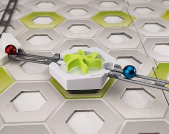 Pode incluir: Um jogo de corrida de bolas com uma bola vermelha e uma azul numa pista cinzenta. A pista está montada numa base de azulejos hexagonais com detalhes verdes e brancos. Um elemento de plástico verde está no centro.