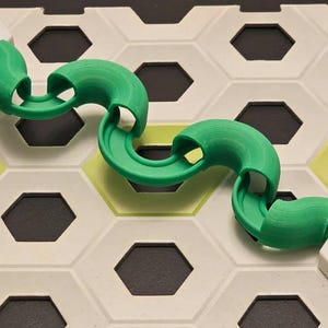 Puede incluir: Una pieza de pista de plástico verde brillante y curvada para una pista de canicas o un juego similar. La pista está colocada sobre una base de baldosas hexagonales con detalles en blanco, negro y amarillo-verdoso. La pista tiene un diseño suave y redondeado.