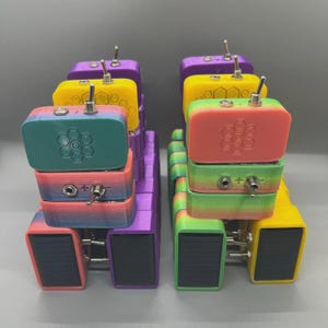 May include: A collection of colorful, stacked electronic devices. Each device features a rectangular shape with a toggle switch, and a honeycomb pattern. The devices are in various colors, including purple, yellow, green, and rainbow gradients.