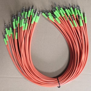 Pode incluir: Um feixe de cabos de áudio laranja com pontas verdes e conectores prateados. Os cabos estão dispostos em forma curva, presos por uma abraçadeira preta. O fundo é uma superfície castanho-claro.