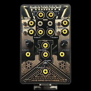 Puede incluir: Un módulo procesador audiovisual Passive Video Toolbox en negro y dorado de Hive Mind Synthesis. El dispositivo rectangular cuenta con múltiples conectores RCA amarillos de entrada y salida, interruptores y perillas giratorias etiquetadas.