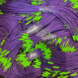 Puede incluir: Un montón de cables de audio morados con puntas verdes brillantes. Los cables están agrupados y envueltos en plástico transparente. Los conectores son plateados. La imagen es una toma de primer plano de los cables.