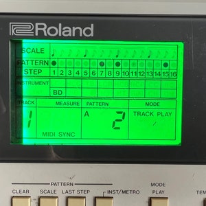 Op de afbeelding: Een groen scherm op een Roland-muziek sampler toont het woord "SCALE" en een muzikaal nootpatroon. Het scherm toont ook het woord "PATTERN" met een zwarte stip, het woord "STEP" met de nummers 1 tot en met 16, het woord "INSTRUMENT" met het woord "BD" eronder, het woord "TRACK" met het nummer "1" eronder, het woord "MEASURE" met de letter "A" eronder, het woord "PATTERN" met het nummer "2" eronder en het woord "MODE" met de woorden "TRACK PLAY" eronder. Het scherm toont ook de woorden "MIDI SYNC".