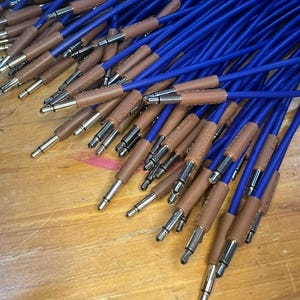 May include: A collection of blue audio cables with brown leather-like sleeves and silver connectors. The cables are arranged on a light brown wooden surface, creating a visually appealing display of audio equipment.