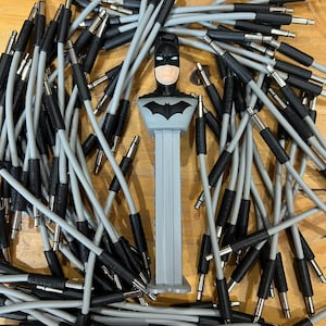 May include: A Batman PEZ dispenser stands amidst a pile of gray and black audio cables. The PEZ dispenser features a black Batman mask and chest emblem. The cables have silver connectors and are scattered across a wooden surface.
