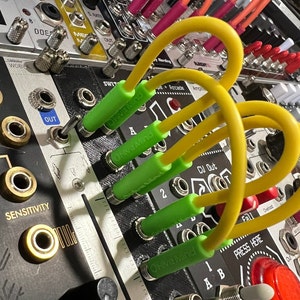 Op de afbeelding: Close-up van vier groene en gele patchkabels aangesloten op een synthesizermodule. De kabels zijn gelabeld "Olive Mind" en de module heeft een knop met het label "Sensitivity".