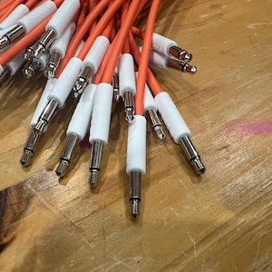 Puede incluir: Un conjunto de cables de audio naranjas con conectores blancos y plateados. Los cables están dispuestos sobre una superficie de madera, mostrando los enchufes y el color vibrante de los cables.