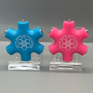 May include: Two audio splitters, one blue and one pink, each with six ports. The text "HIVE MIND SYNTHESIS" is printed on each splitter. The splitters are on clear plastic stands.