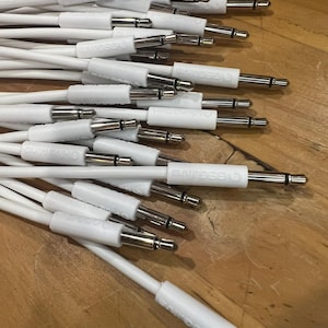 May include: A collection of white audio cables with silver-colored connectors. The cables have white plastic housings with text printed on them. The connectors are silver and black. The cables are lying on a wooden surface.