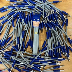 May include: A Batman-themed PEZ dispenser stands amidst a pile of gray audio cables with blue connectors. The PEZ dispenser features a blue cowl and a light-colored face. The cables are scattered on a wooden surface.