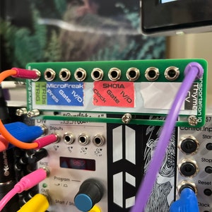 May include: A close-up of a green circuit board with multiple jacks and labels, including "MicroFreak", "SHOIA", "TRIG", "Gate", "1V/0", "Clock", "CV", and "Thyme".  The board is connected to a synthesizer with various knobs, buttons, and jacks.