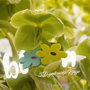 Könnte beinhalten: Nahaufnahme einer grünen Pflanze mit großen Blättern. Zwei blütenförmige Ringe sind abgebildet, einer türkis und einer gelb, mit einem schwarzen Zentrum. Das Wort "bloom" ist weiß überlagert, zusammen mit dem Text "3D gedruckte Ringe".