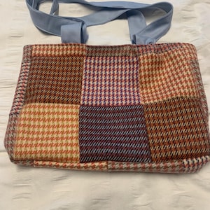 May include: A patchwork tote bag with a pale blue handle. The bag's body is made of various tweed squares in shades of brown, red, and purple. The bag is rectangular and appears to be made of a soft, woven material.
