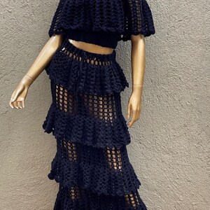 Peut inclure: Un ensemble deux pi&egrave;ces en crochet noir compos&eacute; d'un haut court &agrave; &eacute;paules d&eacute;nud&eacute;es avec des manches &agrave; volants et d'une jupe longue assortie &agrave; &eacute;tages. Le v&ecirc;tement pr&eacute;sente un motif en maille ajour&eacute;e cr&eacute;ant un aspect textur&eacute; semi-transparent.